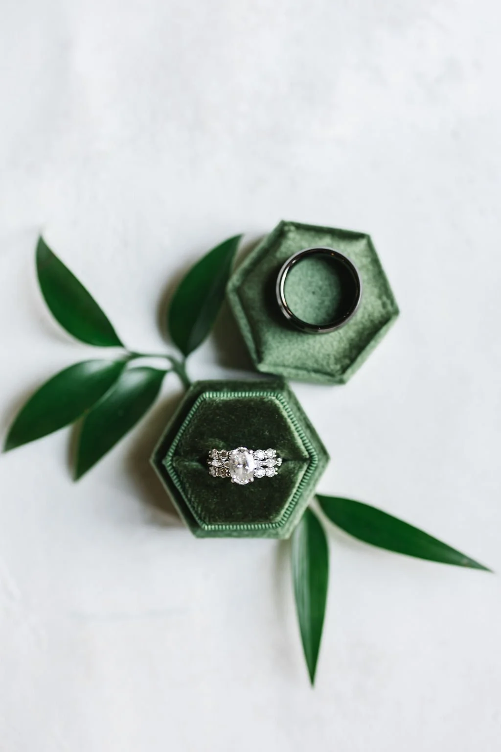 A jewelry ring with diamonds inside a green hexagonal velvet box, with a black ring and green leaves on a white background.