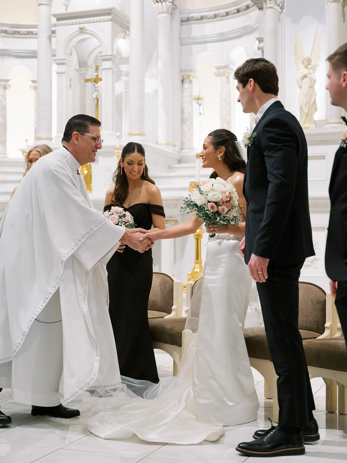 A wedding ceremony taking place in a church, with a bride and groom exchanging vows. The priest is holding the bride's hand, and the bride is smiling while holding a bouquet of white and pink roses. Bridesmaids and groomsmen are standing beside them 