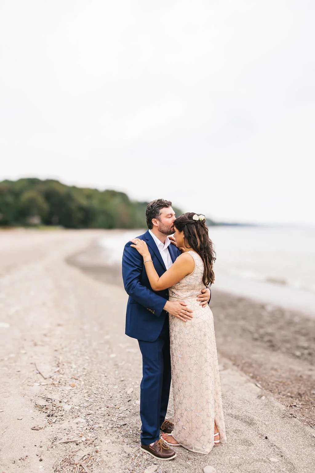 A couple dressed in formal wedding attire embracing on a beach, with the man in a navy suit and the woman in a long lace dress with a floral hair accessory, on a cloudy day.