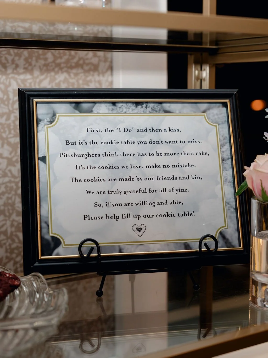 A framed sign with a poem about cookies and a request for donations, displayed on a table with a glass of water and pink flowers to the right.