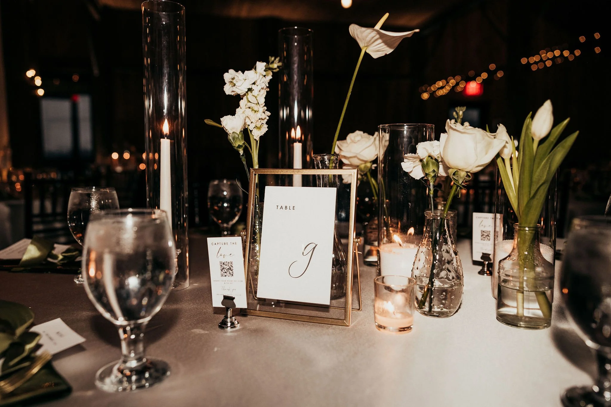 Elegant table centerpiece with white flowers in glass vases, surrounded by wine glasses, candles, and a framed table number with the letter 'g', set for an event or celebration.