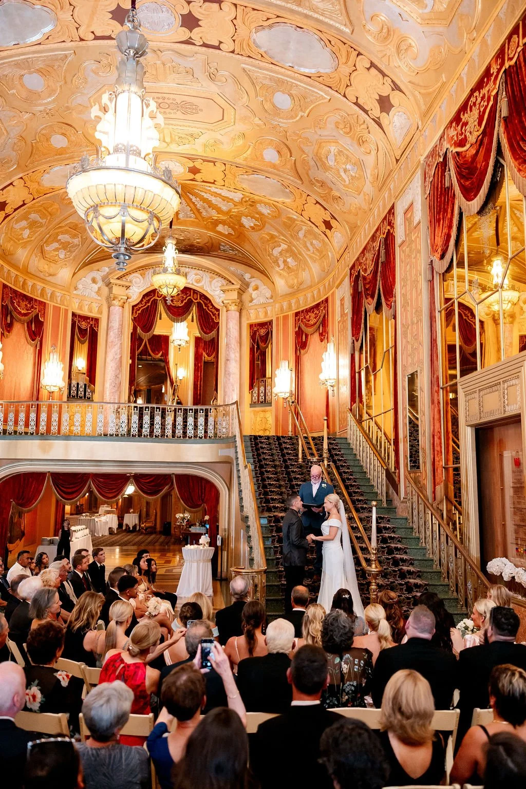 A wedding ceremony taking place in a grand, ornately decorated hall with high ceilings, chandeliers, and red velvet curtains. The bride and groom are standing at the base of a staircase, exchanging vows in front of an officiant, while guests watch an