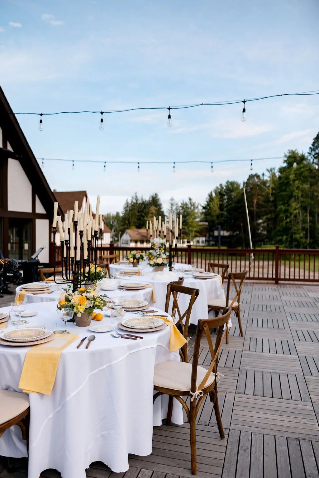 Outdoor dining setup with round tables covered in white tablecloths, decorated with floral centerpieces and candles, set for a formal event on a wooden deck with string lights overhead in a scenic area with trees and houses.