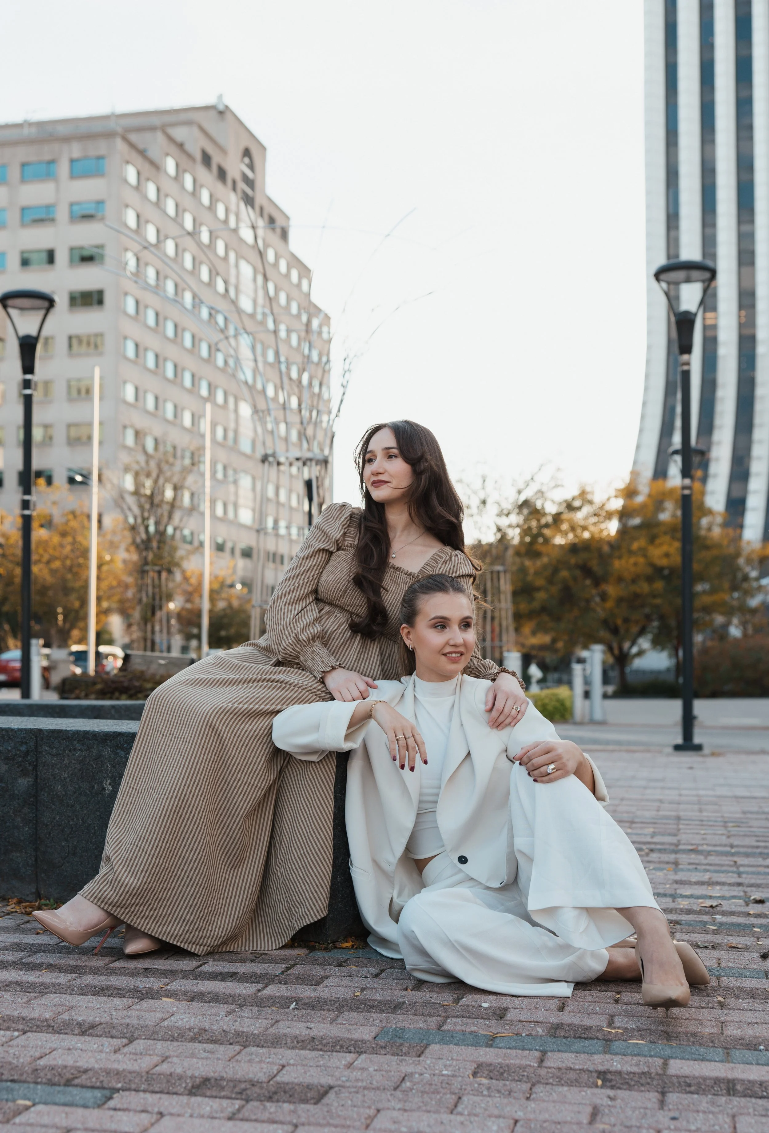 Two women posing outdoors in an urban setting with tall buildings and streetlights, one sitting on the ground and the other standing behind her. They are dressed in stylish outfits, with autumn trees in the background.