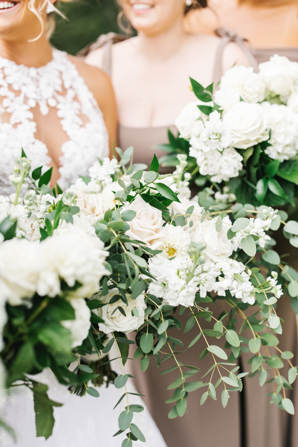 Close-up of white floral bouquet with greenery, with two women in the background, one in a lace wedding dress and the other in a beige dress, smiling at a wedding.