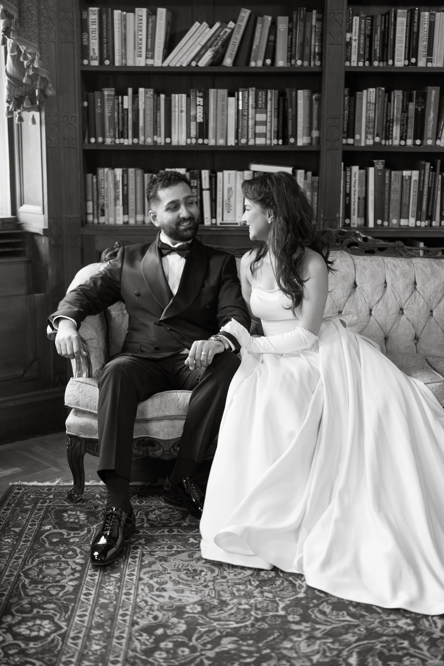 A black and white photo of a man and woman sitting on an ornate sofa in a library. The man is dressed in a tuxedo and the woman in a wedding dress. They are holding hands and smiling at each other.