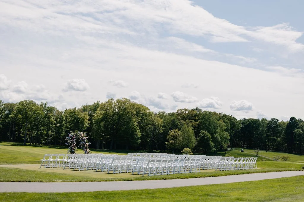 Outdoor wedding ceremony setup with white chairs arranged in rows facing a floral arch, on a grassy field with trees in the background and a partly cloudy sky overhead.