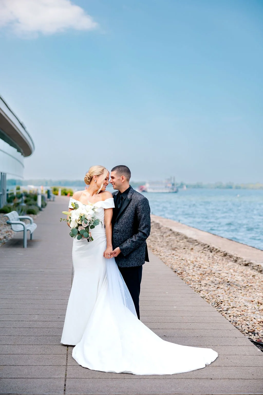 A bride and groom happily holding hands by a waterfront on their wedding day, with a clear blue sky in the background.