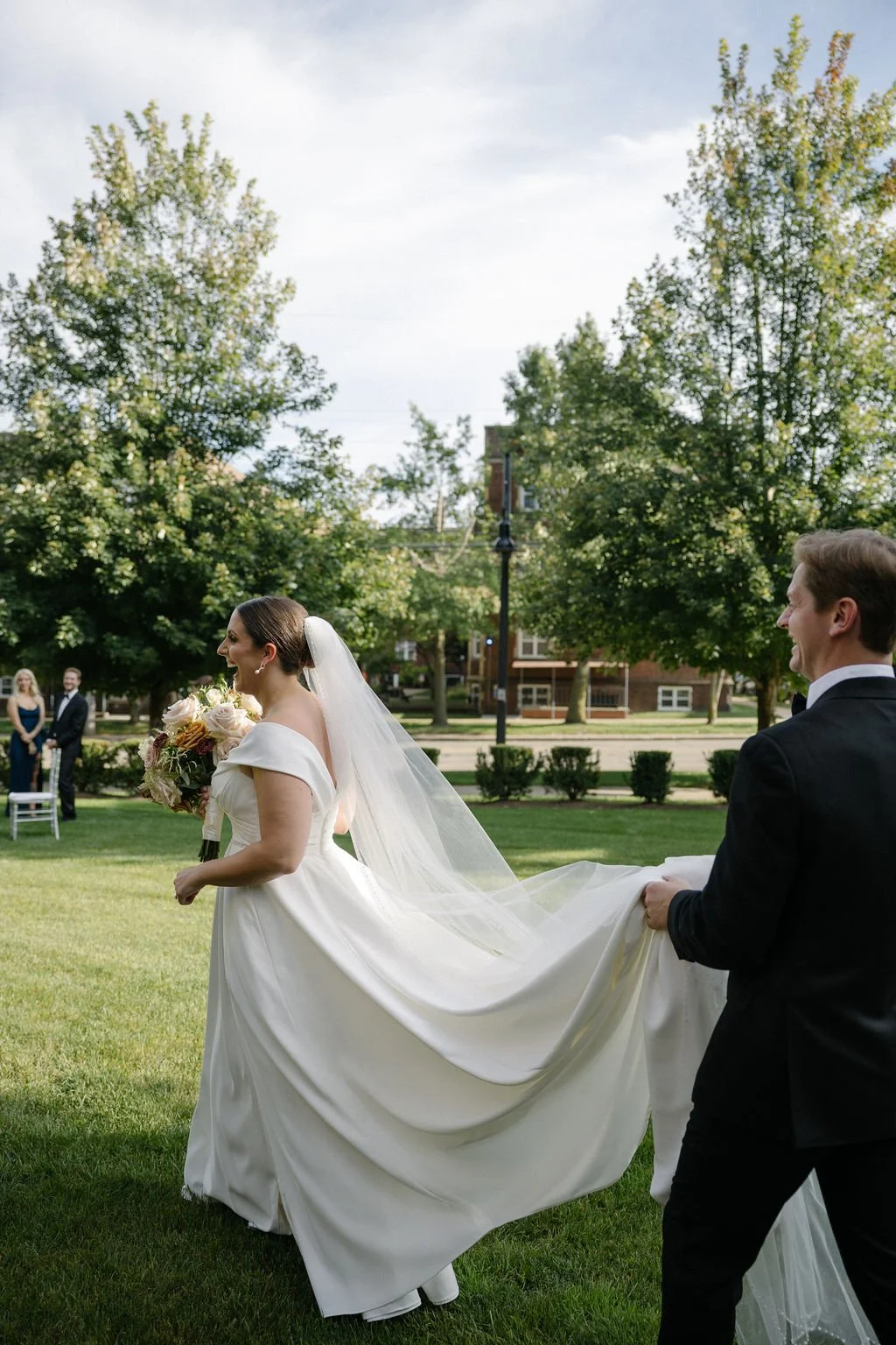 Bride in a white wedding dress and veil holding a bouquet, walking on grass with groom holding her train, outdoor wedding ceremony with guests in the background and green trees.