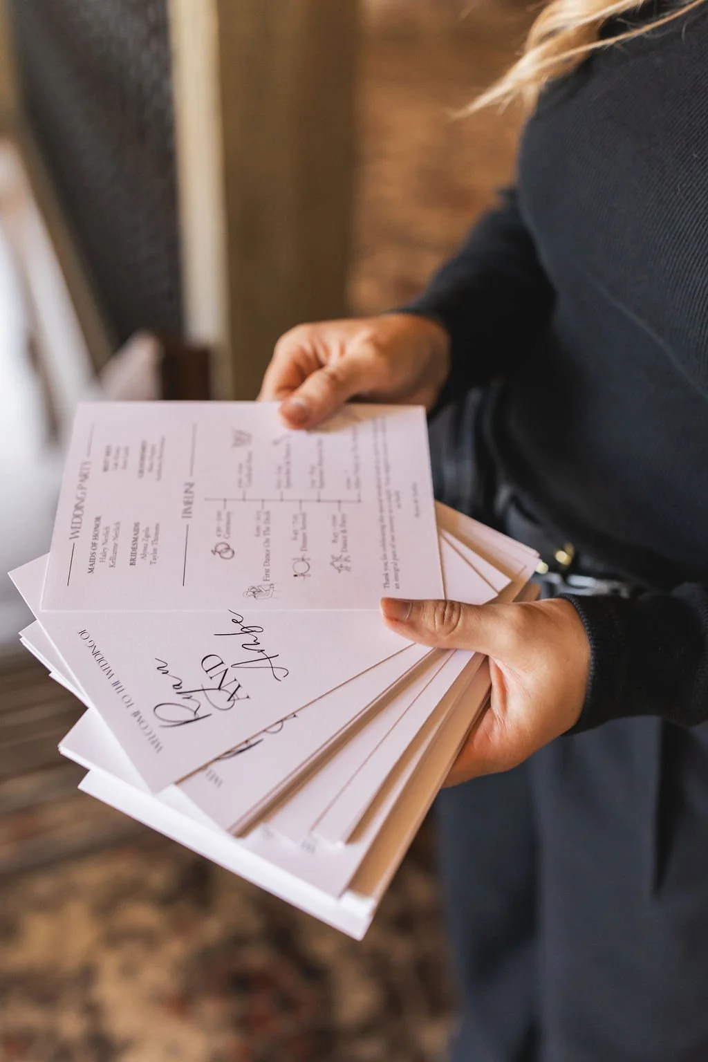 Person holding a stack of wedding programs or menus, dressed in dark clothing, with wooden flooring in the background.