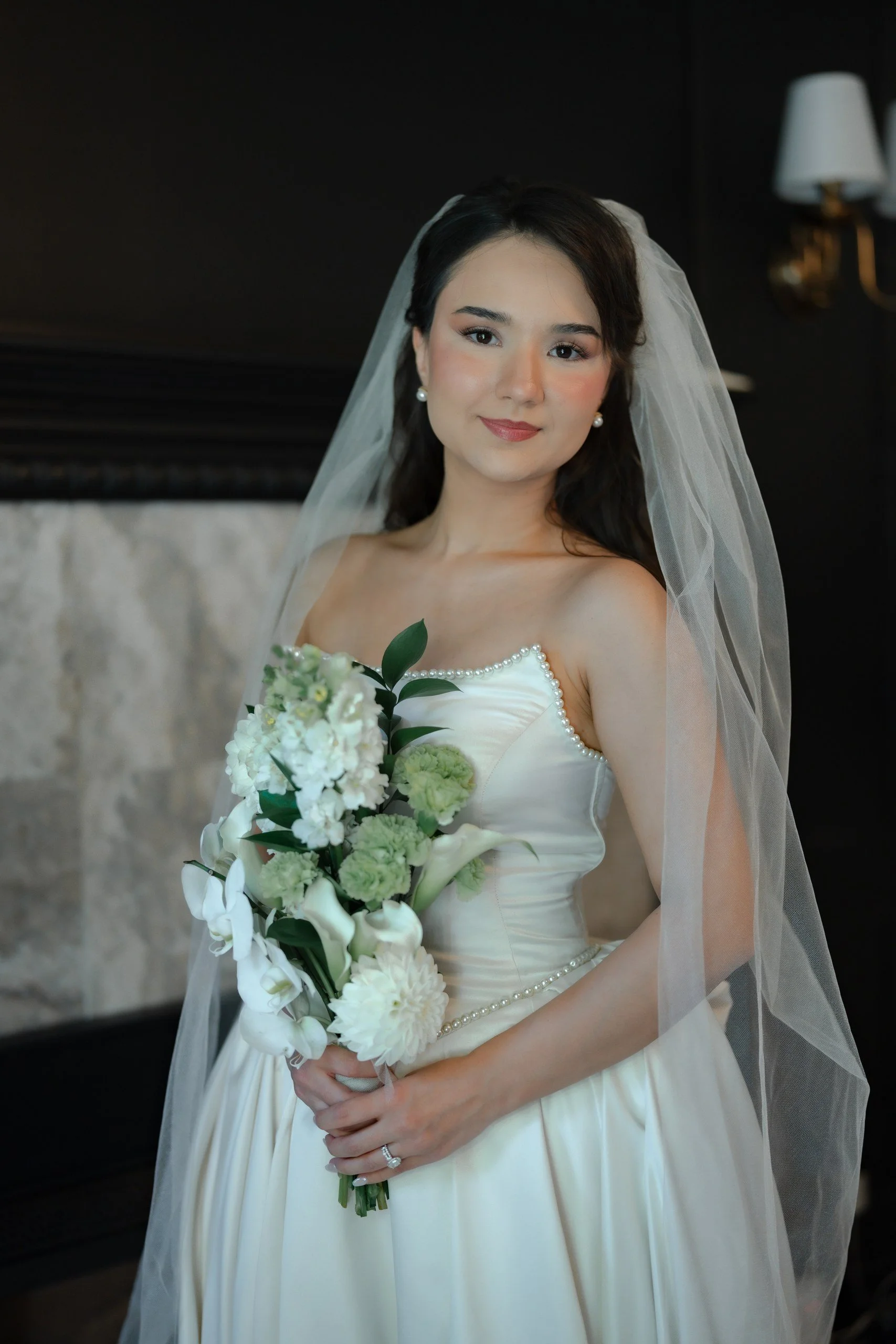 A young woman in a wedding dress holding a bouquet of white and green flowers, wearing a veil and pearl earrings, standing indoors with a dark wall and light fixtures in the background.