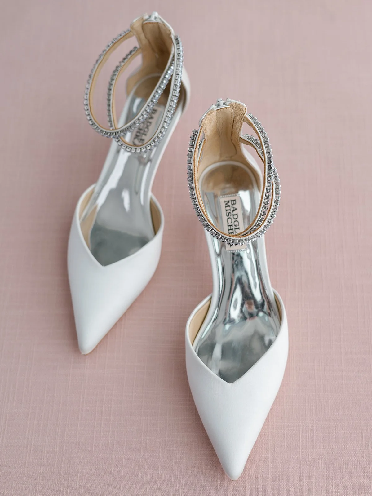 White high-heeled shoes with pointed toes and clear insoles, decorated with rhinestone ankle straps, placed on a pink surface.