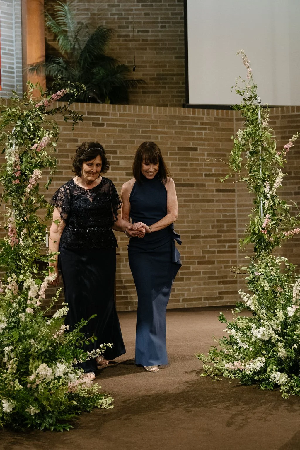 Two women walking hand in hand through a floral arch at a formal event.