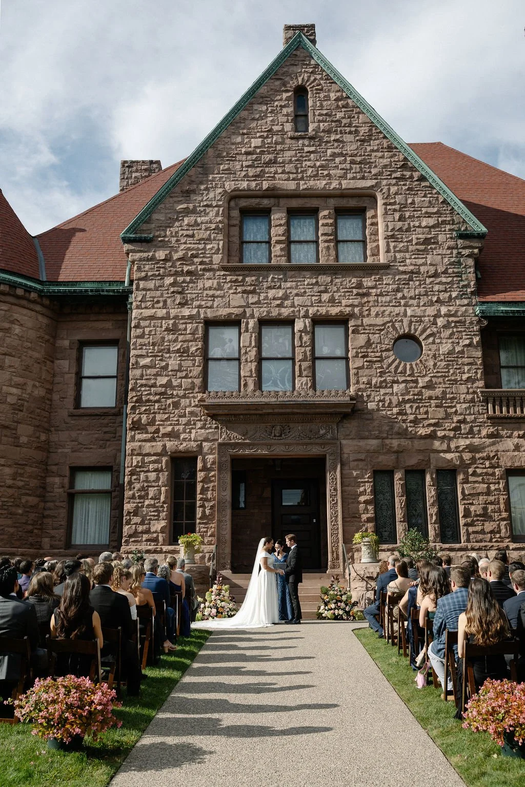 A wedding ceremony outdoors in front of a large stone building with Gothic architecture. Bride and groom stand facing each other, holding hands, while guests sit on either side of an aisle. Flower arrangements are placed at the steps, and the buildin
