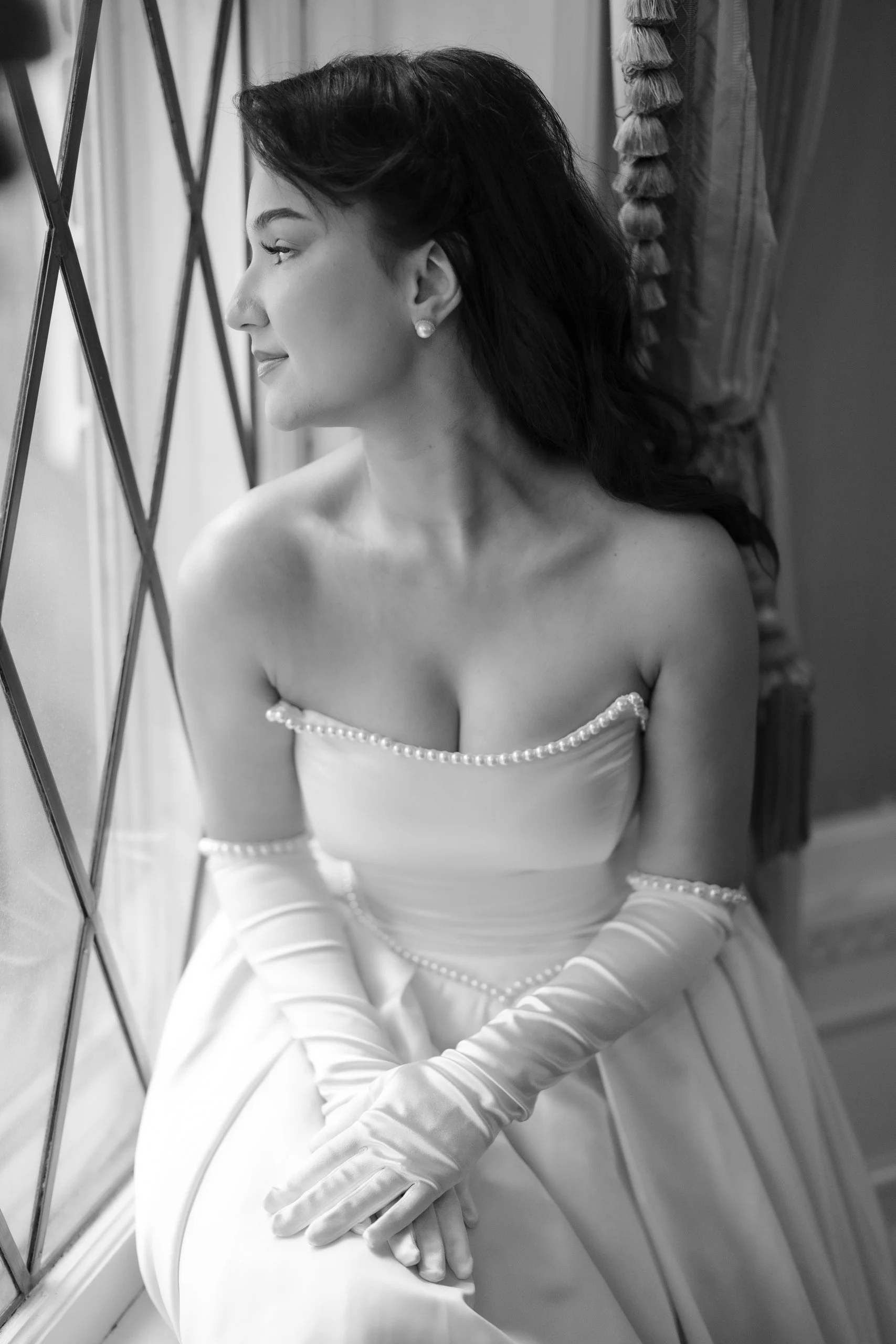 Black and white photo of a woman in a wedding dress, sitting by a window with a geometric pattern. She has dark hair styled back and is wearing pearl earrings and gloves. She appears to be looking out the window with a gentle smile.