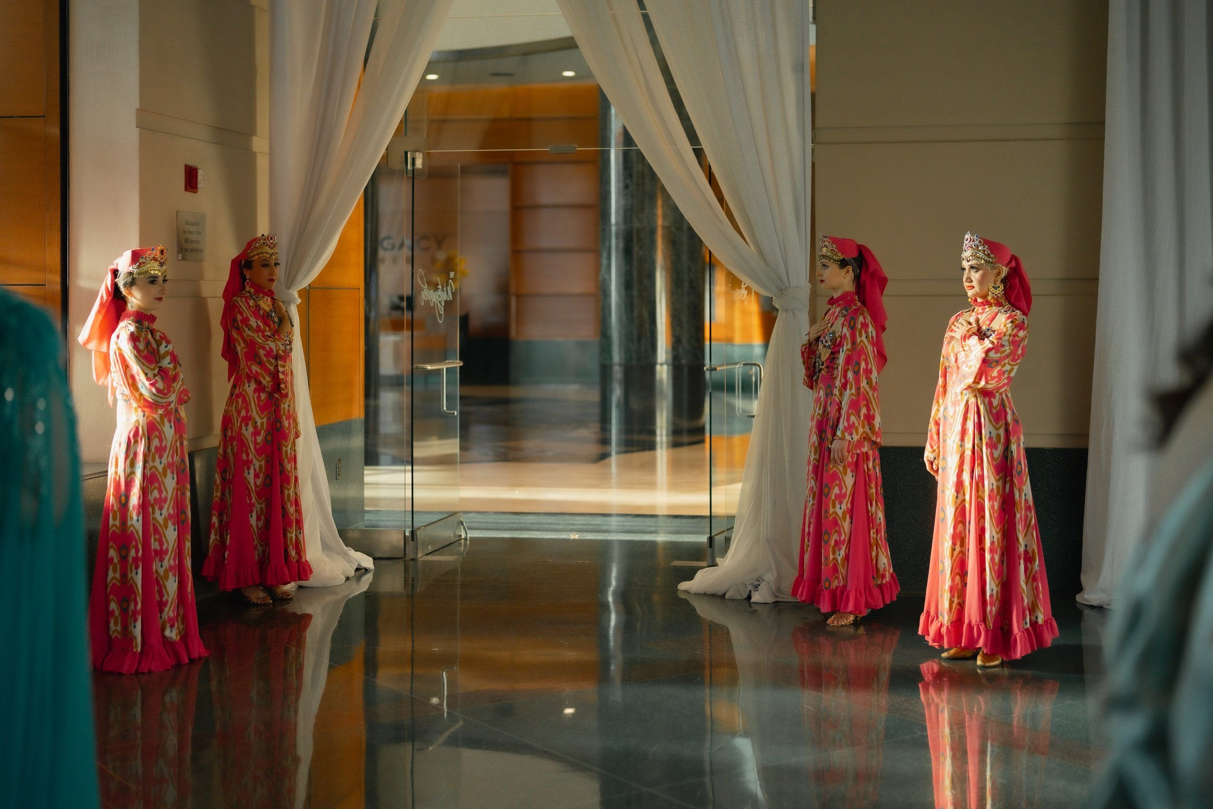Four women in vibrant, patterned dresses and traditional headdresses standing on either side of a white curtain doorway in an indoor setting.