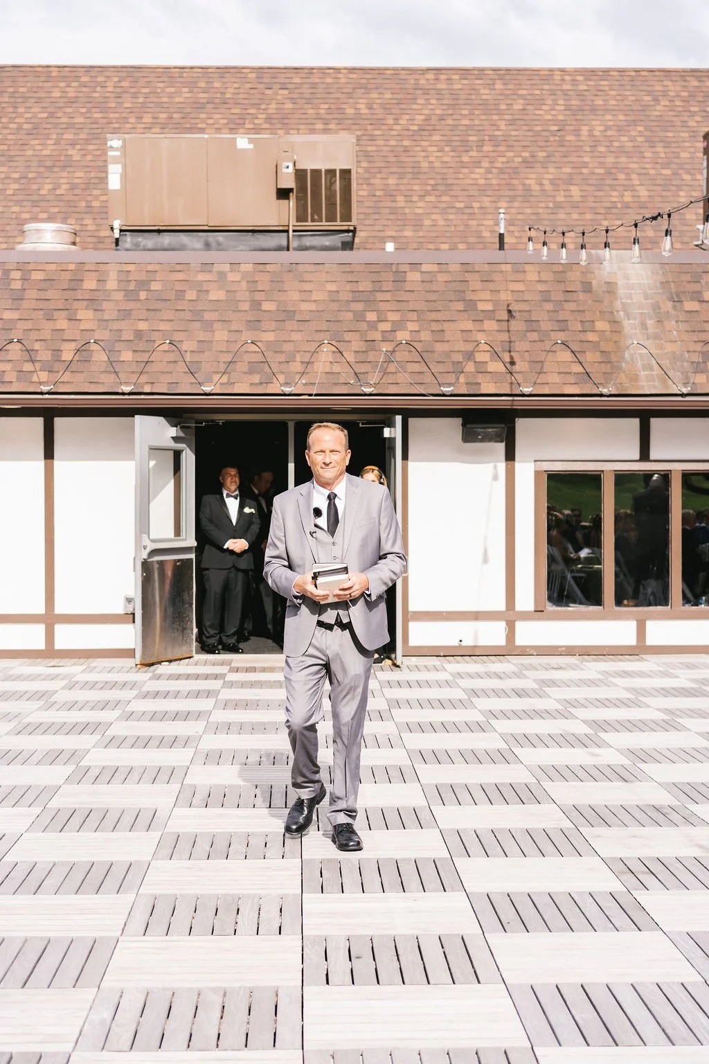 Man in gray suit holding a book, standing outdoors in front of building with people inside, dressed formally, at an event or gathering.