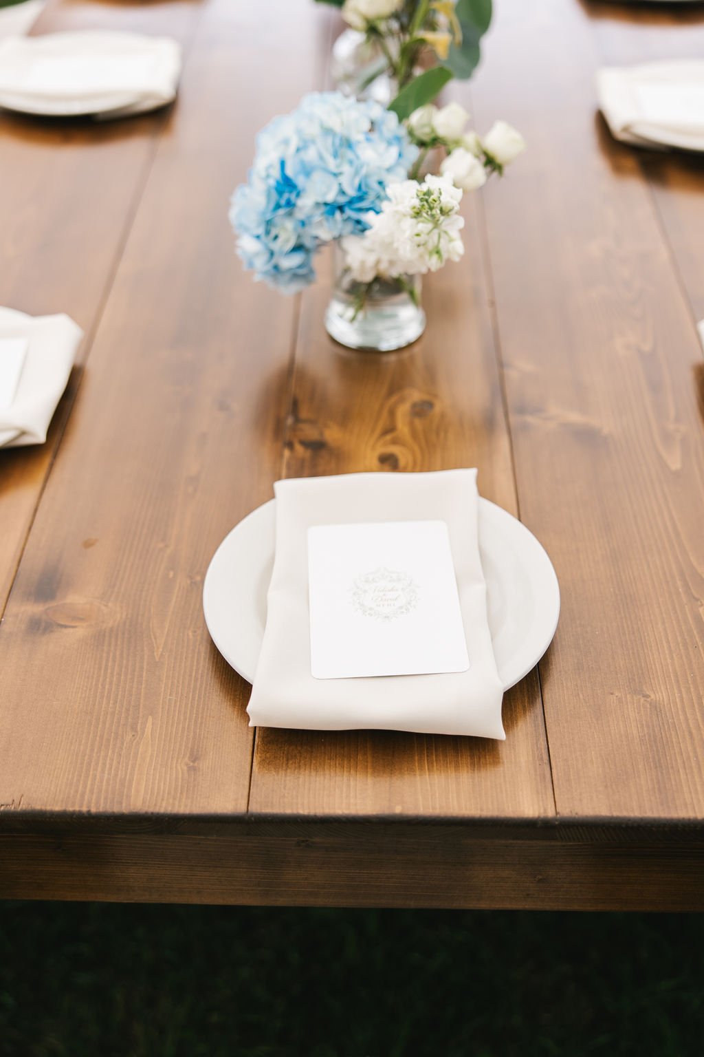 A wooden table decorated with a vase of blue and white flowers, a white plate with a napkin and a menu or card placed on top.