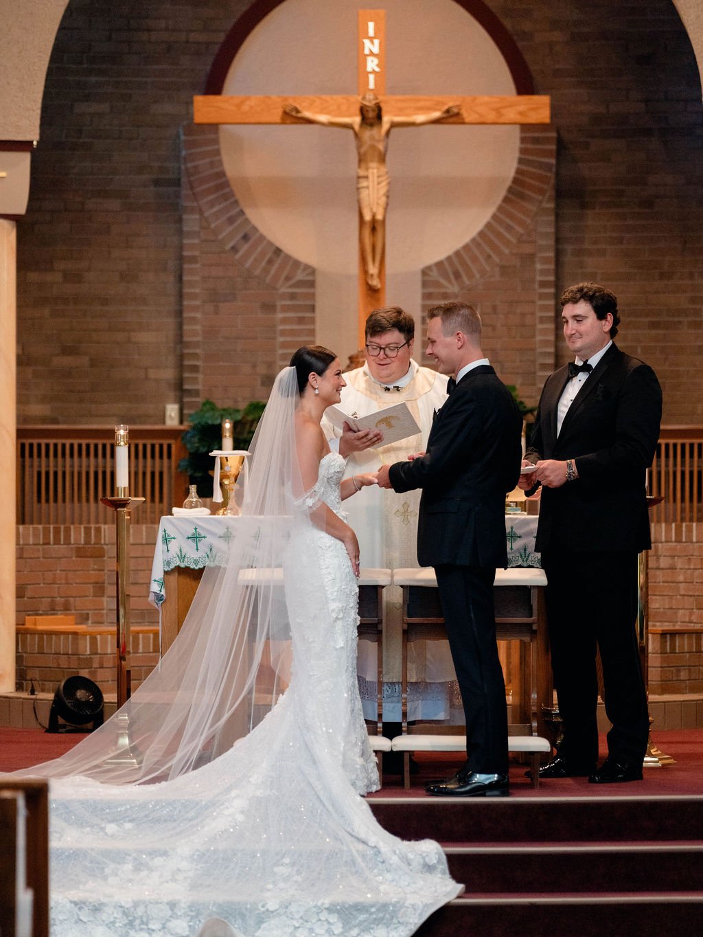 A bride and groom exchange vows at the altar in a church during their wedding ceremony, with a priest officiating and a large crucifix hanging behind them.
