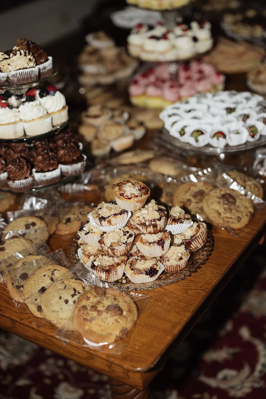 Assorted cookies and cupcakes on a wooden table at a dessert buffet including chocolate chip cookies, crumb-topped berry bars, and cupcakes with whipped frosting and berries.