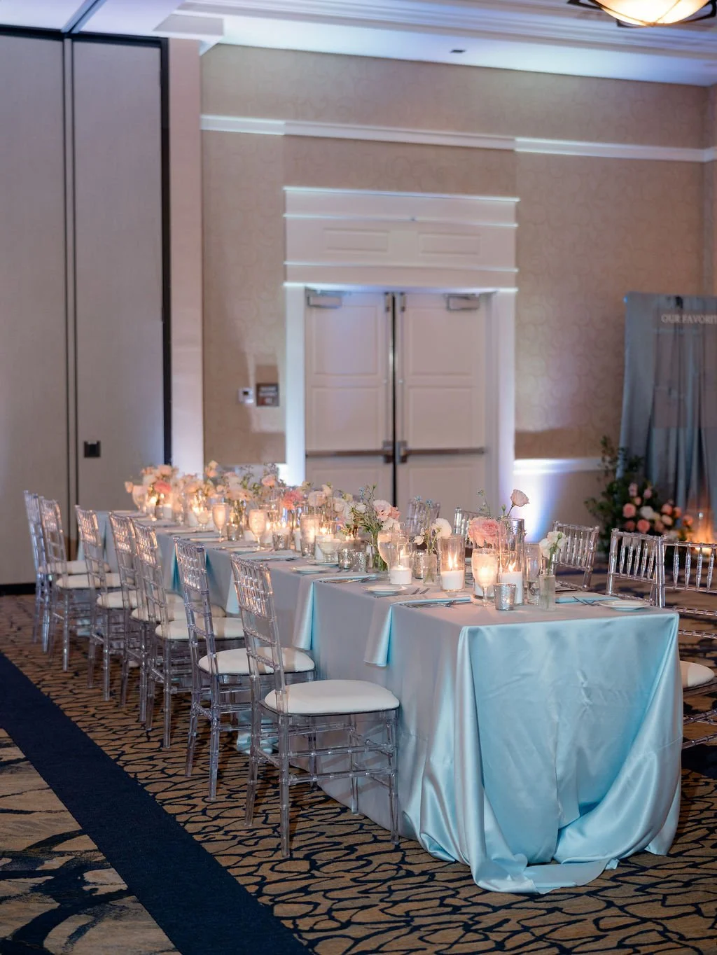 Elegant banquet table set with candles, flowers, and glassware in a decorated event room with seating and floral arrangements.