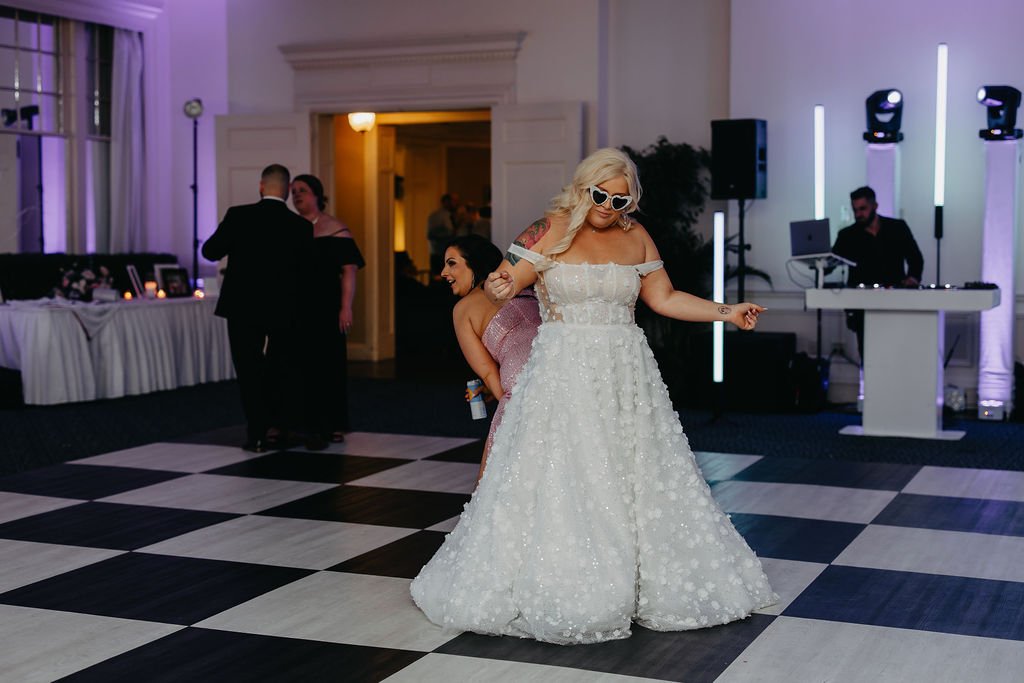 Woman in a white wedding dress and sunglasses dancing on a checkered dance floor at a wedding reception, with a DJ playing music in the background and other guests in the background.