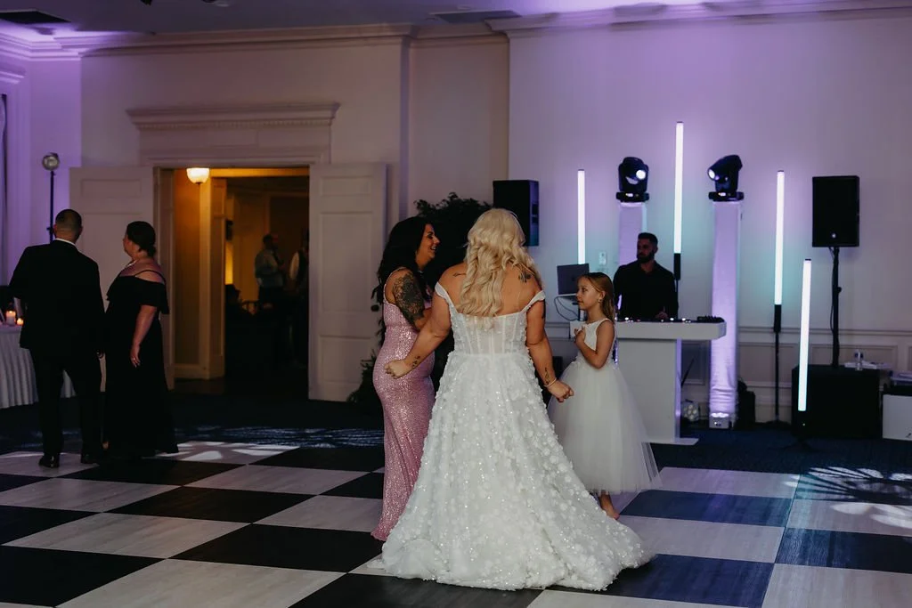 People dancing and socializing at a wedding reception on a checkered dance floor, with a DJ booth and colorful lighting in the background.