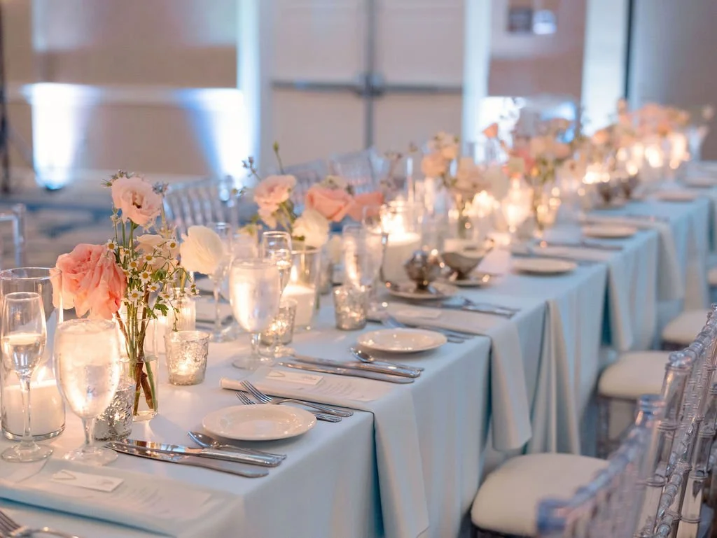 Long reception table decorated with pink and white flowers, candles, glassware, and place settings in a well-lit reception hall.