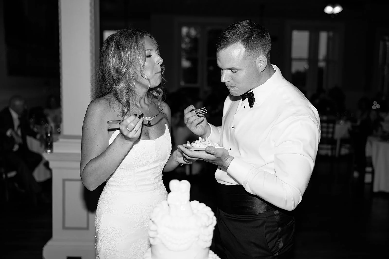 A bride in a white wedding dress and a groom in a tuxedo with a bow tie sharing a slice of wedding cake at their reception.