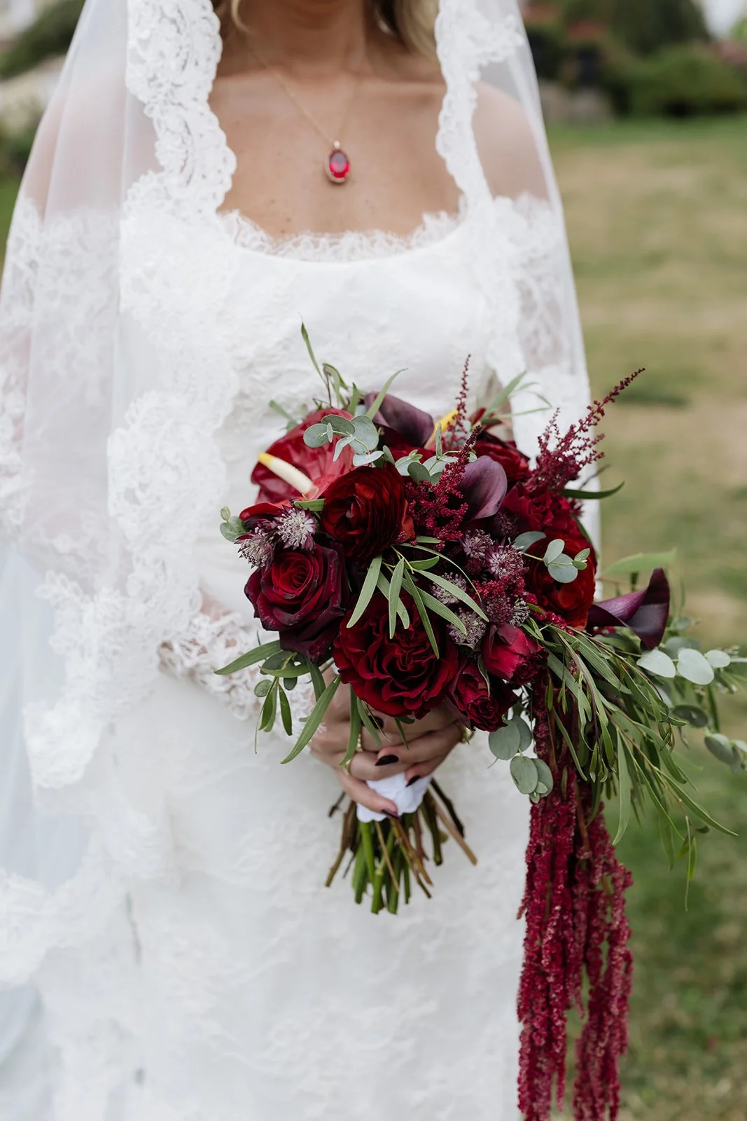 Bride holding a bouquet of deep red roses, calla lilies, and greenery, wearing a lace wedding dress and a necklace with a red pendant, outside on a grassy area.