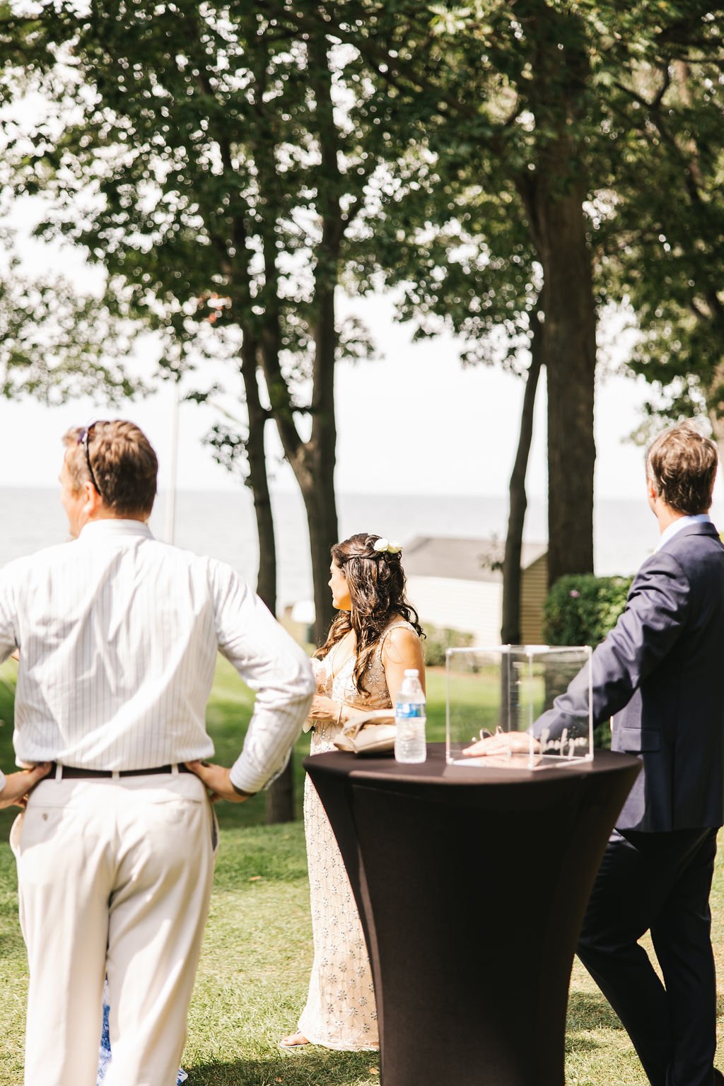 Three people standing outdoors near a table with a water bottle and a glass box, with trees and a house in the background, during daytime.