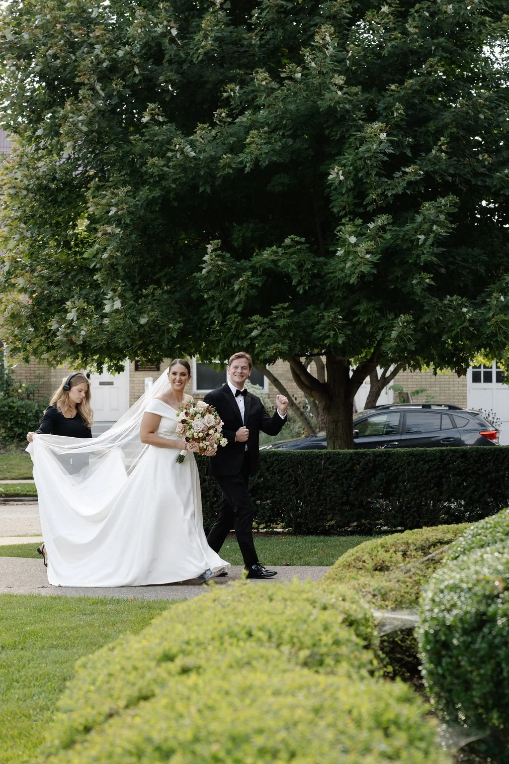 Bride and groom walking outside, smiling, with the bride holding a bouquet of flowers, under a large green tree, with a woman adjusting the bride's dress in the background.