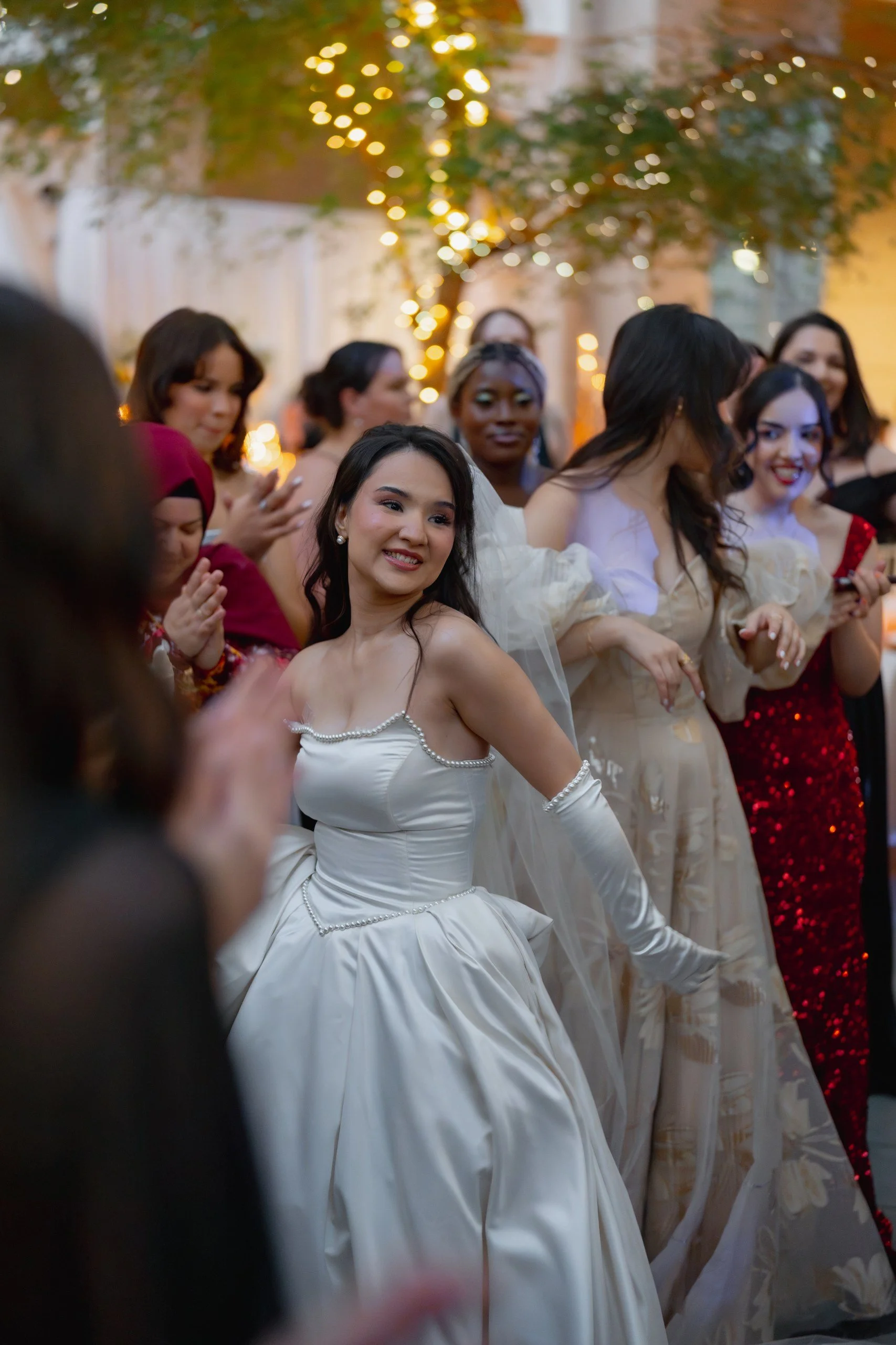 A group of women in elegant dresses attending a celebration or wedding, smiling, surrounded by festive lighting and outdoor decor.