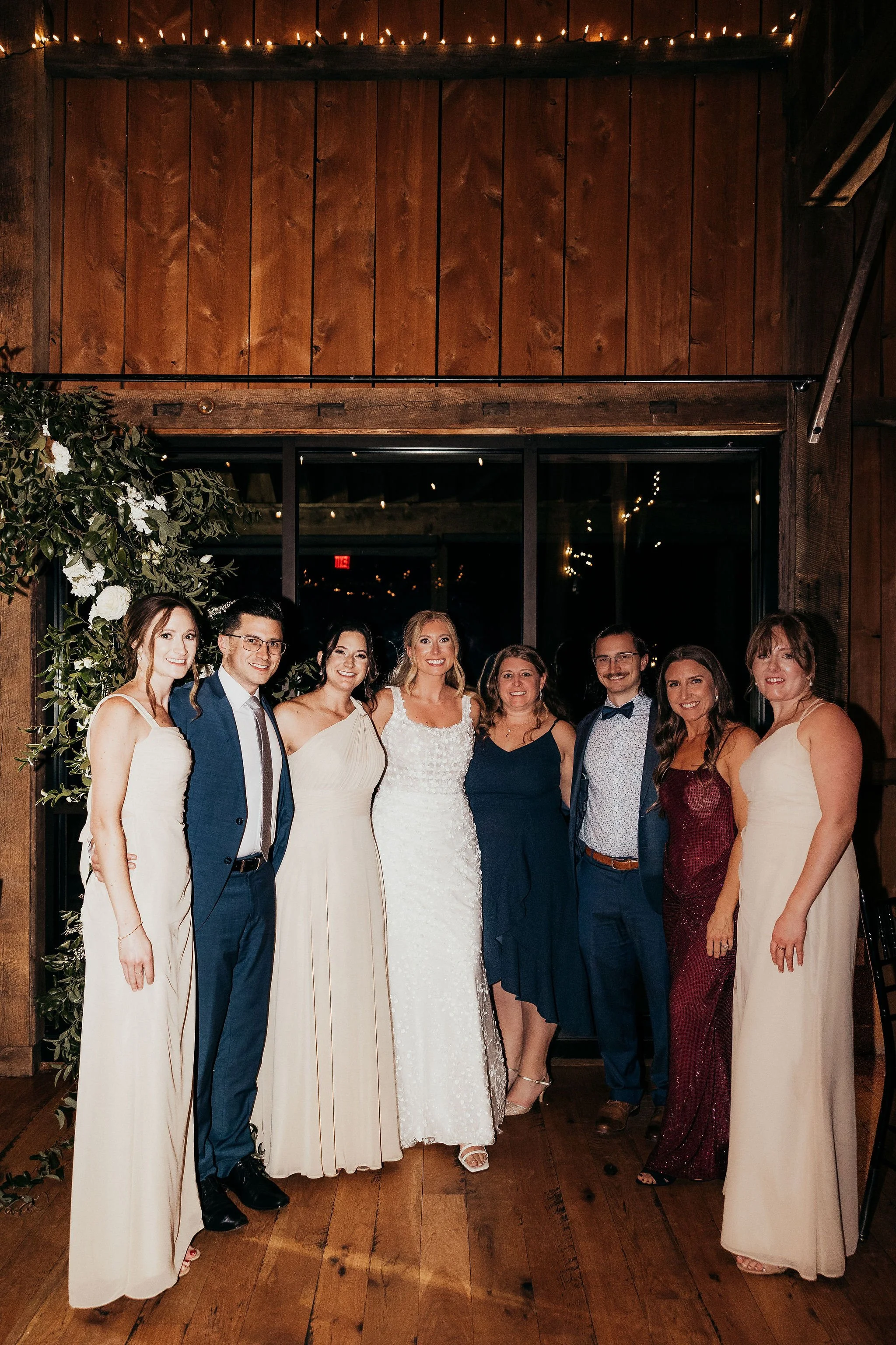 A group of ten well-dressed people posing together indoors at a celebration or wedding, with warm lighting, wooden walls, and a window behind them.