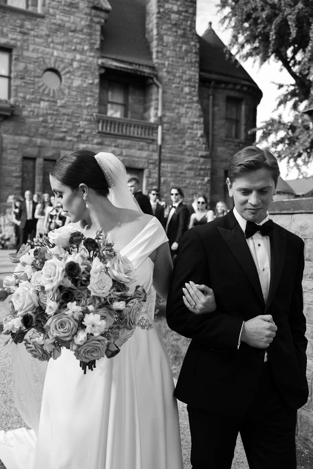 A bride in a white wedding dress and a groom in a black tuxedo, walking arm-in-arm outdoors with a group of people in the background, in front of a stone building.