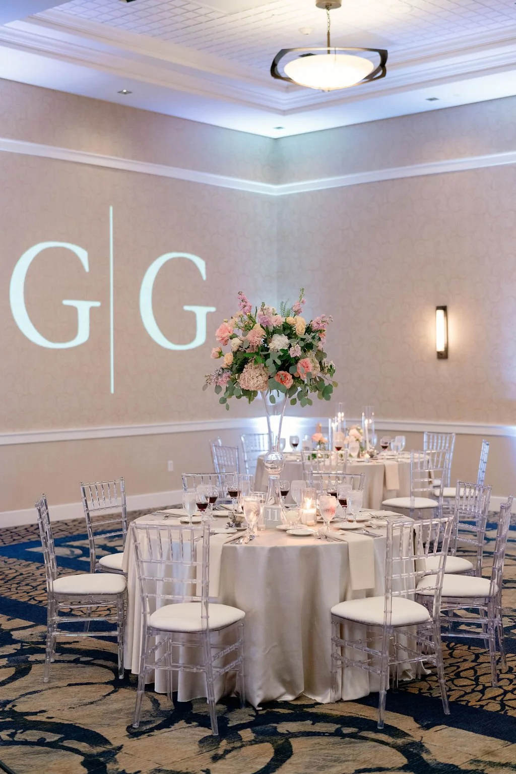 Elegant event space with round tables, white tablecloths, clear chairs, floral centerpieces, and wall projection of initials GG.
