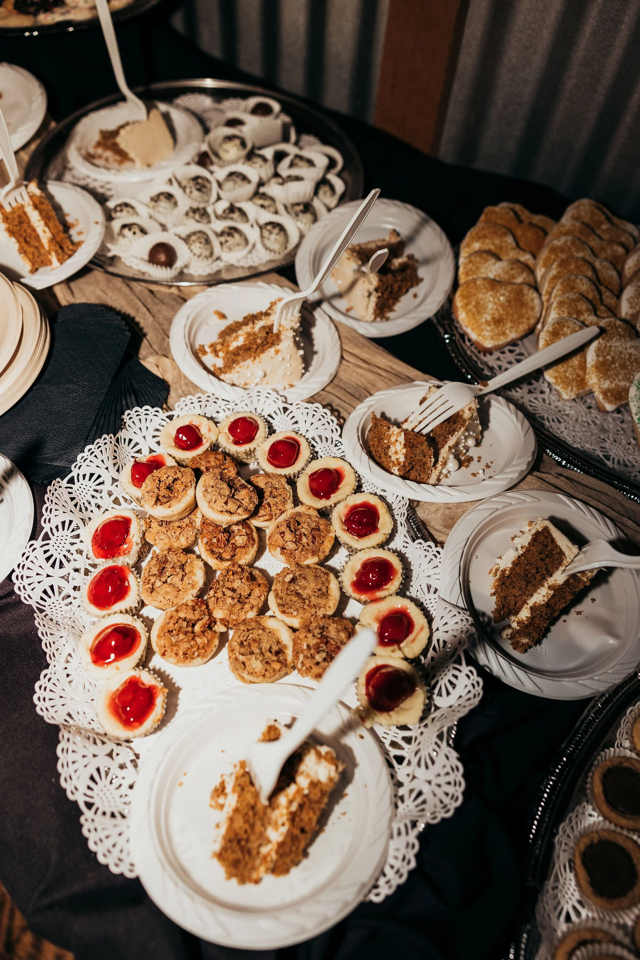 Assorted desserts including slices of cake, cookies, and small pastry cups with cherry toppings arranged on decorative trays and plates at a dessert table.