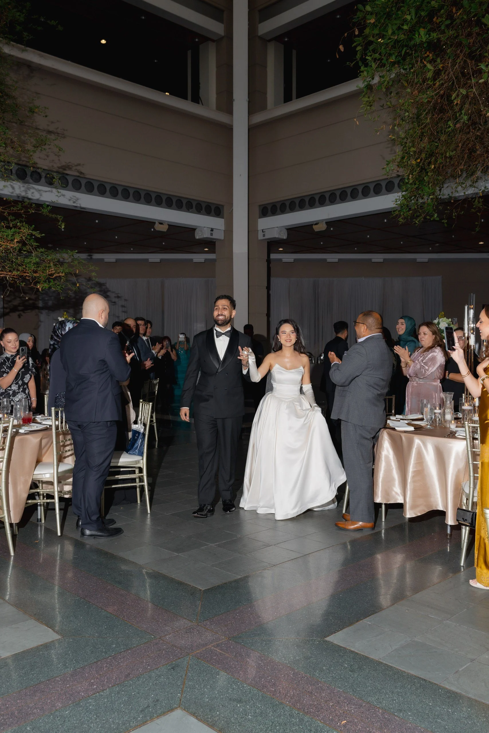 A bride and groom holding hands and smiling at a wedding reception indoors, surrounded by guests clapping and taking photos, with tables and decorations visible.