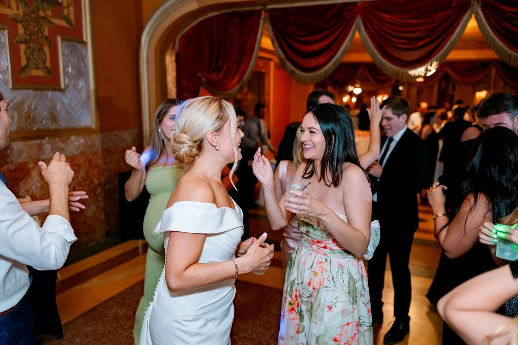People dancing and socializing at a party or wedding reception in an elegant venue with draped curtains and warm lighting.