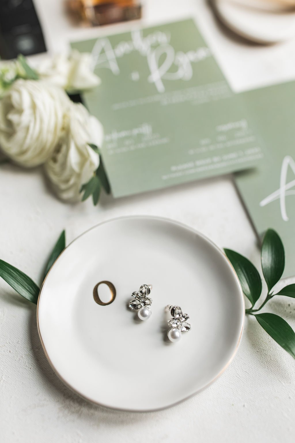 Photo of wedding rings, including a pearl and diamond earrings, on a white plate with green leaves, with a wedding invitation and floral decorations in the background.