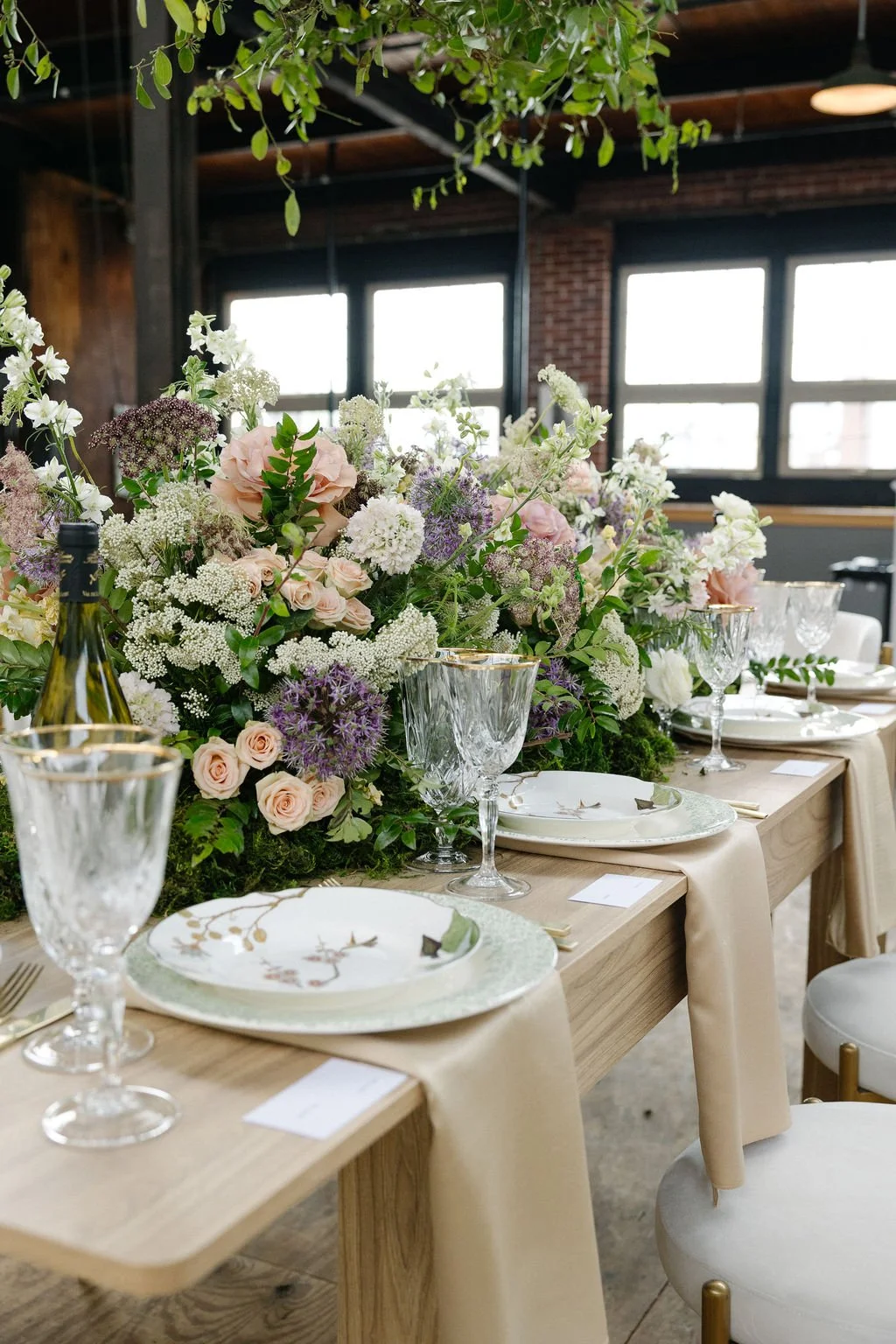 Elegant table setting with floral centerpieces, fine china, crystal glasses, and gold-trimmed plates, arranged on a wooden table in a rustic venue with large windows and greenery hanging from the ceiling.