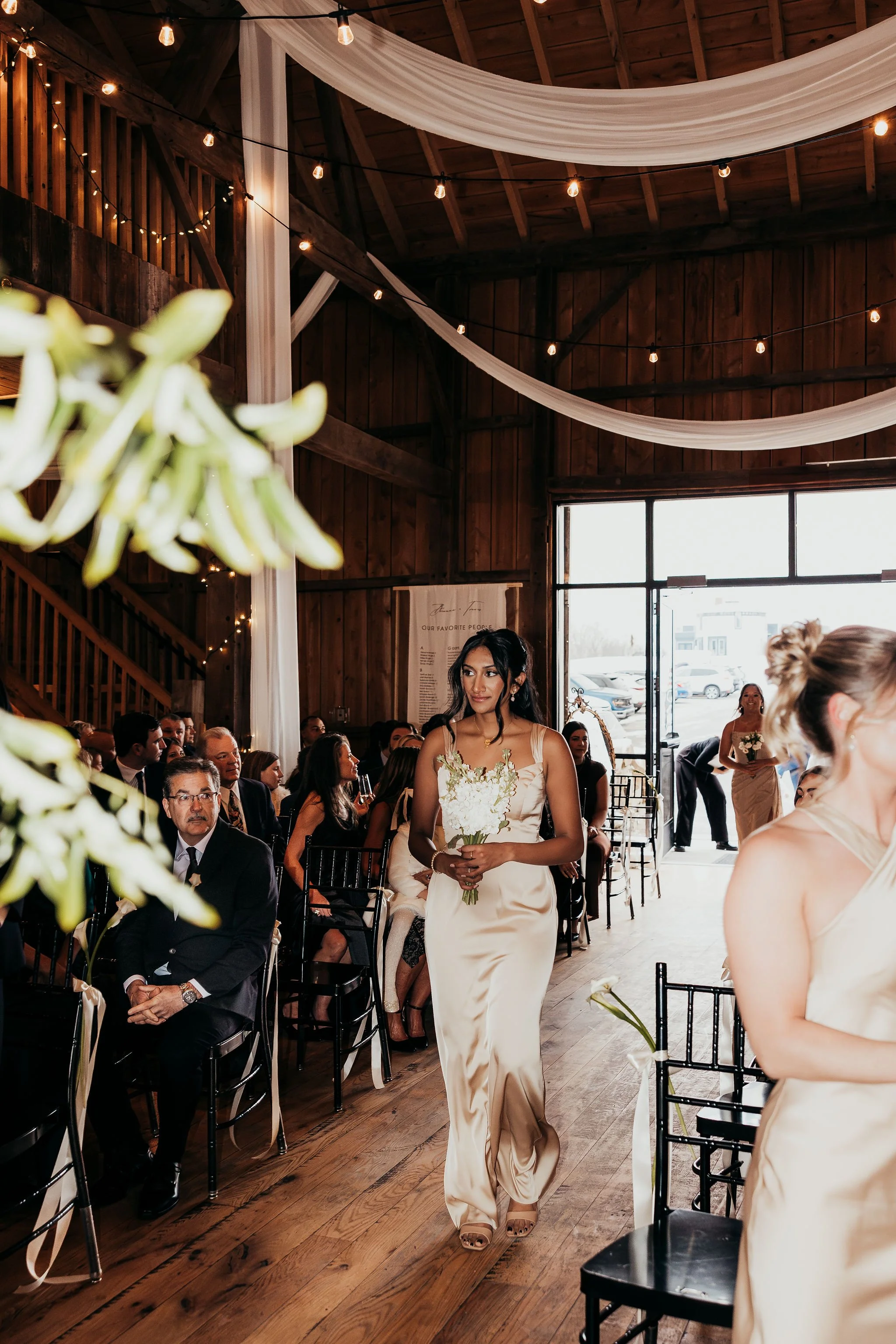 A bride walking down the aisle holding a bouquet at a wedding ceremony inside a rustic wooden venue decorated with draped white fabric and string lights.
