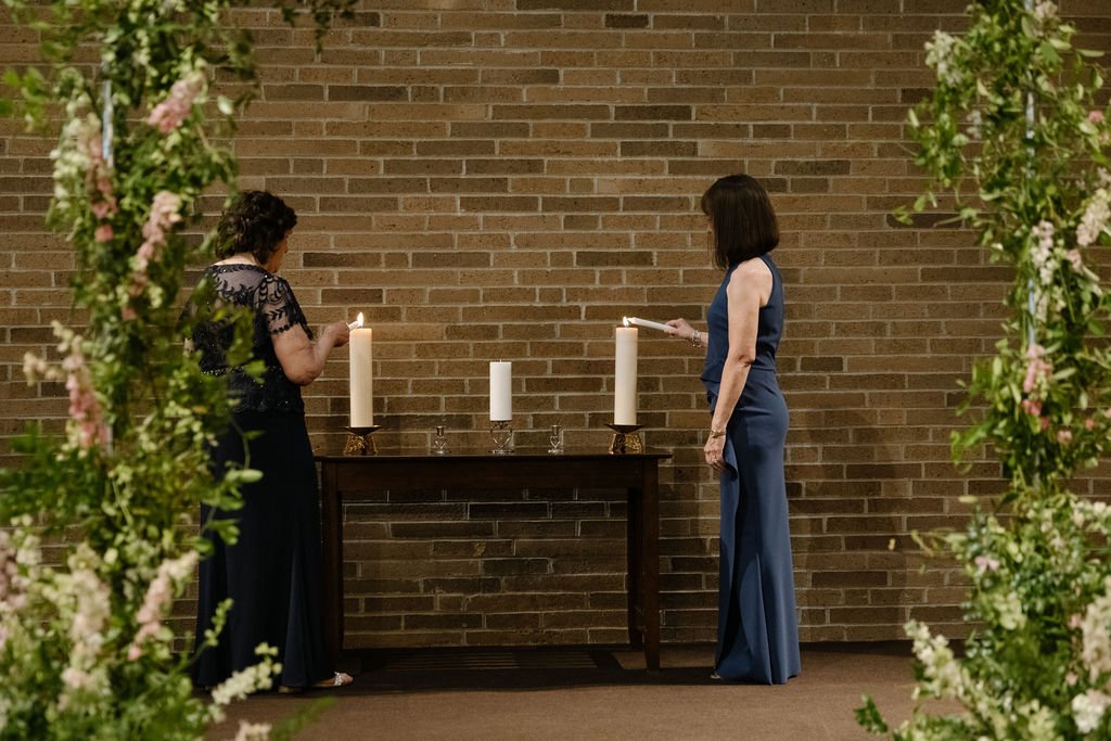 Two women stand on either side of a table with three white candles, lighting them, framed by floral arrangements, against a brick wall background.