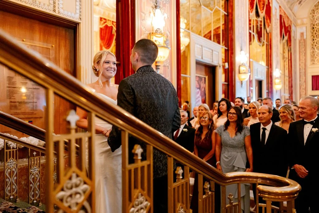 A wedding ceremony taking place in an ornate, luxurious hall with a bride and groom standing at the altar, surrounded by guests dressed in formal attire.