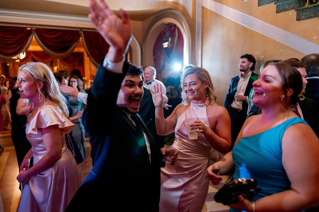 People dancing and celebrating at a formal event or wedding reception in an elegant hall with draped curtains and warm lighting.