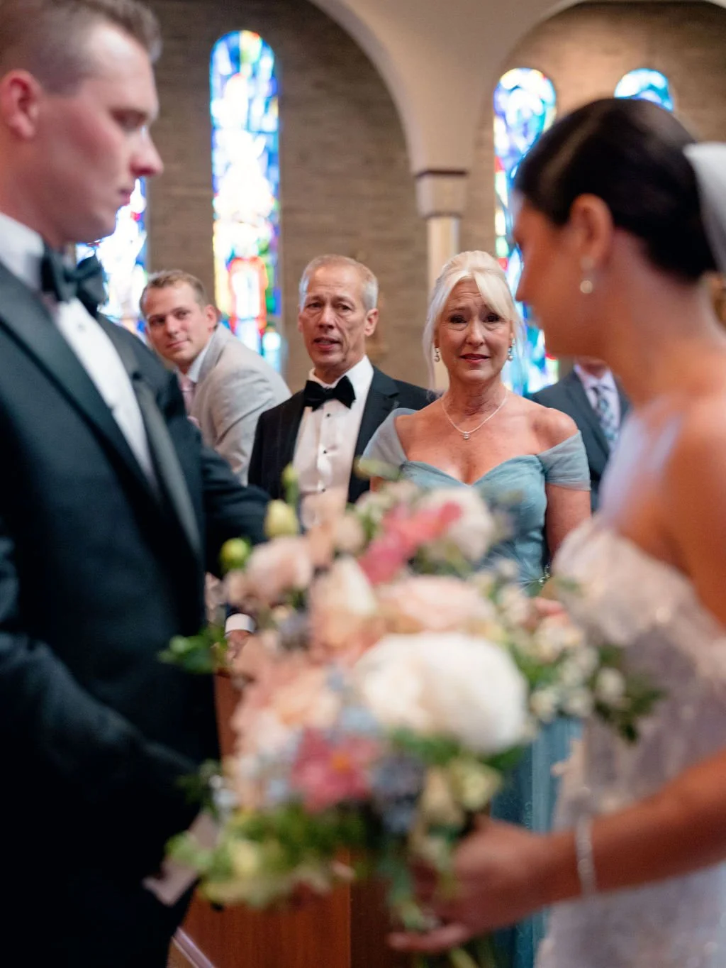 A bride and groom exchanging wedding vows in a church, with close family members watching during the ceremony. The church has stained glass windows in the background.
