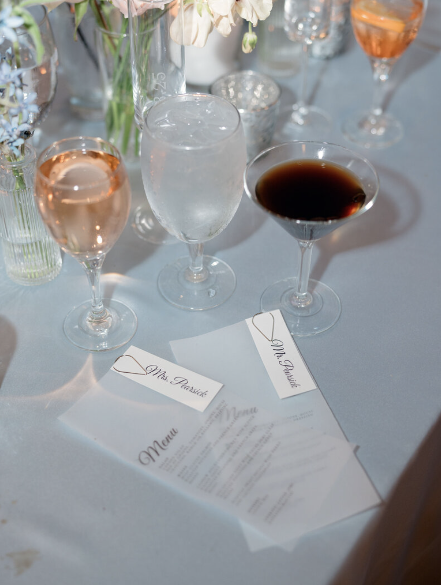 A wedding table setting featuring various glasses with drinks, including a rosé wine, a glass of water, and a dark cocktail. There are flower arrangements in different vases, and a printed menu with place cards labeled 'Mrs. Prasid' and 'Mr. Prasid'.