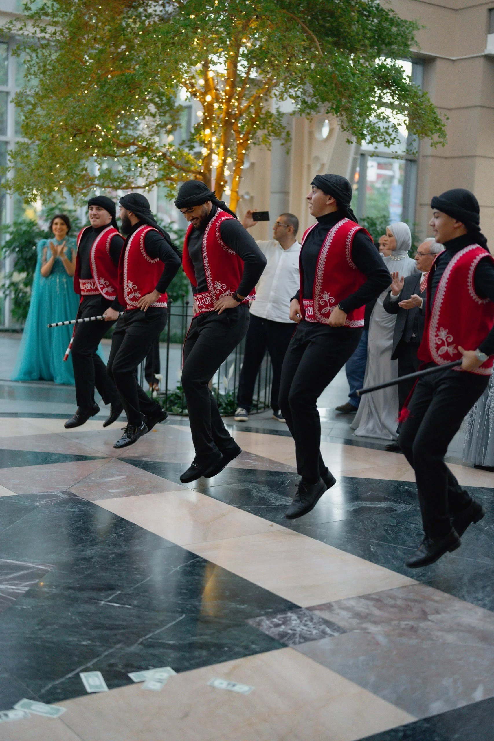 Men dressed in traditional Eastern European costumes dancing indoors, with a woman clapping and other spectators watching in the background.