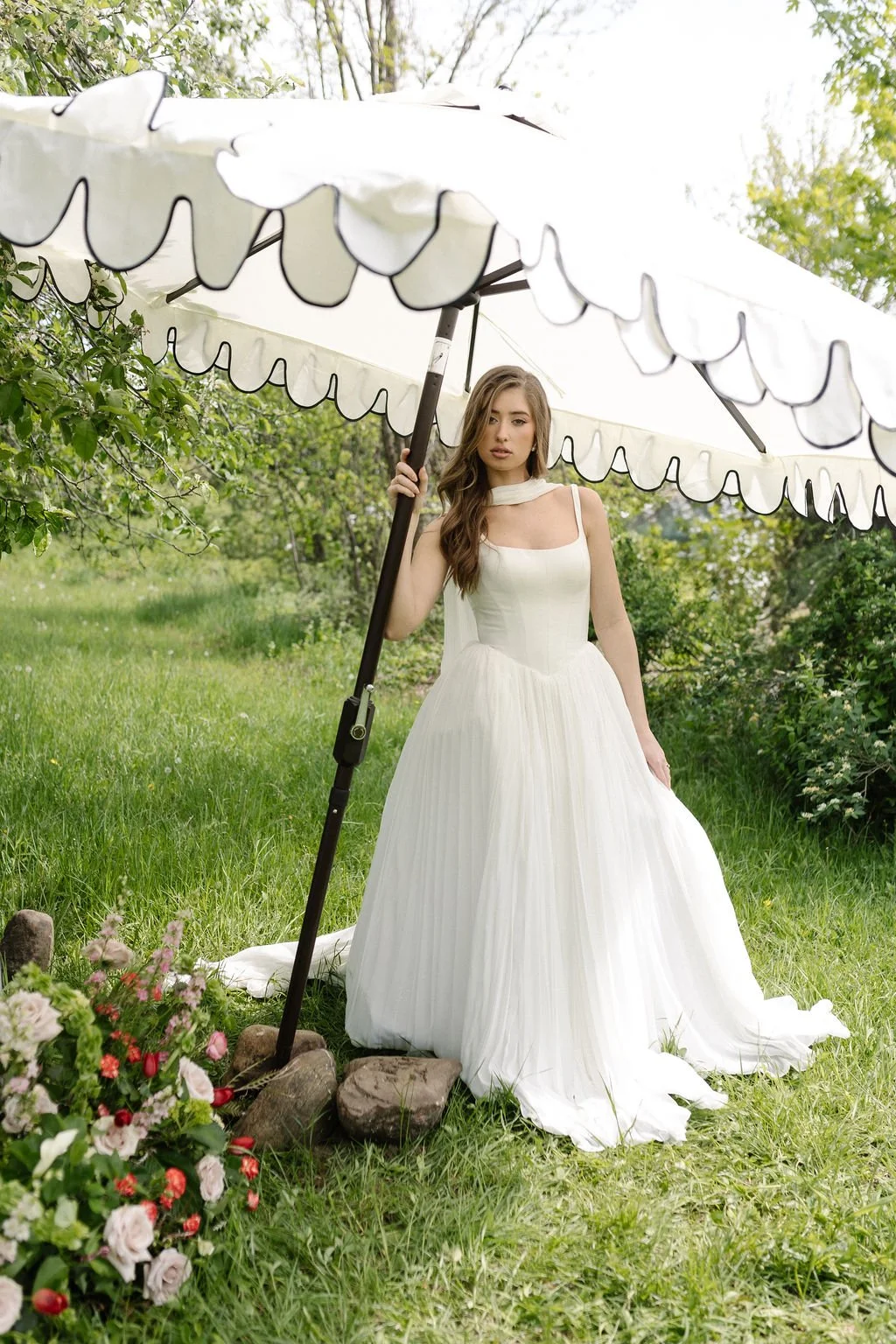 A young woman in a white gown standing outside under a large white umbrella, surrounded by green grass, trees, and flowers.