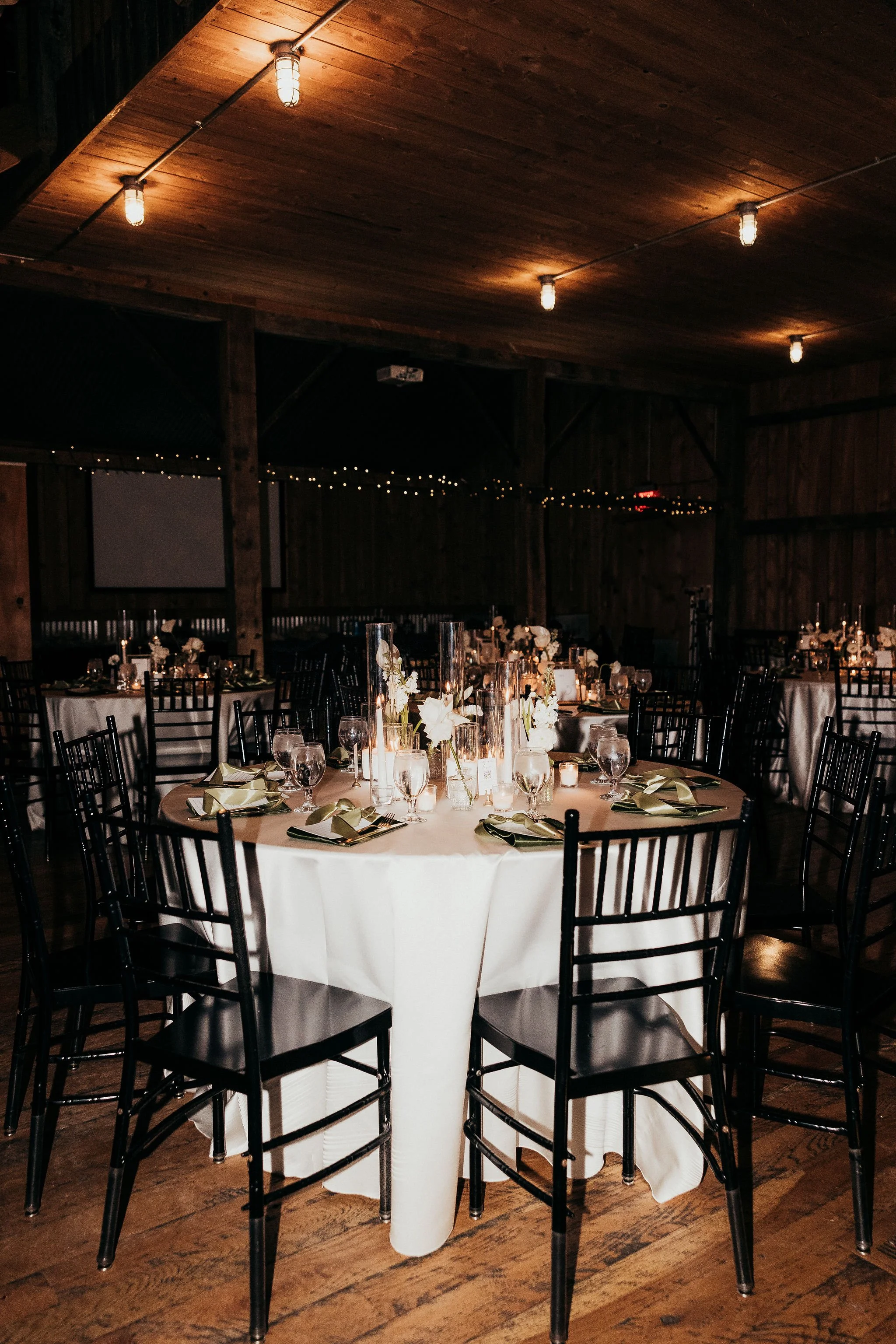 An indoor event space with a round table covered with a white tablecloth, decorated with tall glass vases holding white flowers, surrounded by black chairs, set for a formal dinner, with dim lighting and string lights hanging from a wooden ceiling.
